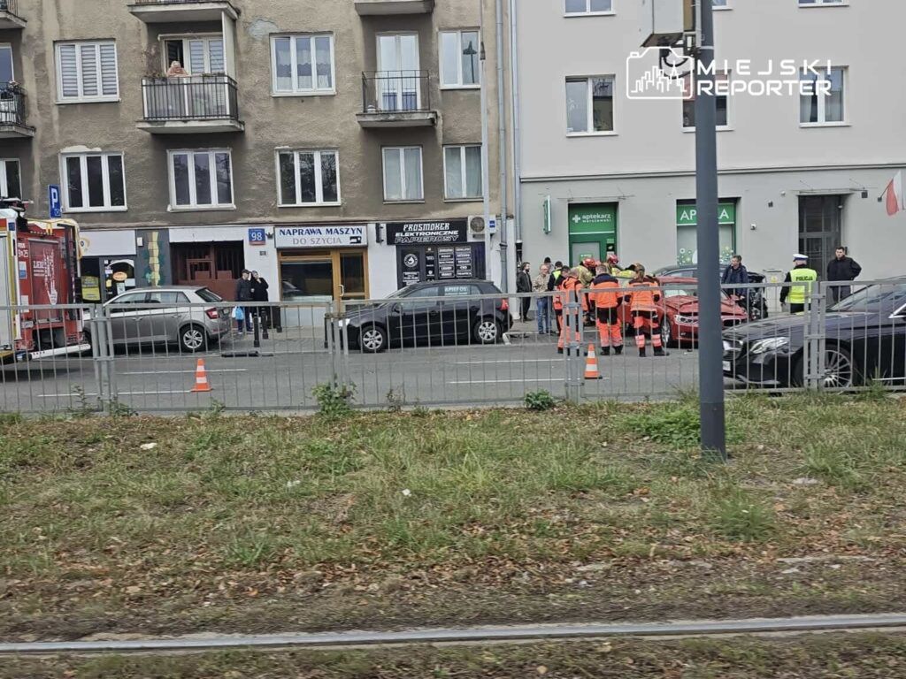 Grupa ratowników w pomarańczowych uniformach rozmawia z policjantem na ulicy, obok wzywającego pomocy samochodu.
