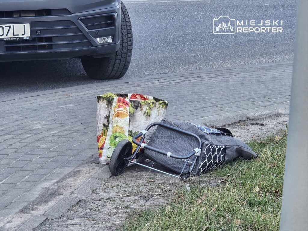 Wózek dziecięcy leży na chodniku obok torby z kolorowym wzorem owoców, w pobliżu zaparkowanego samochodu.