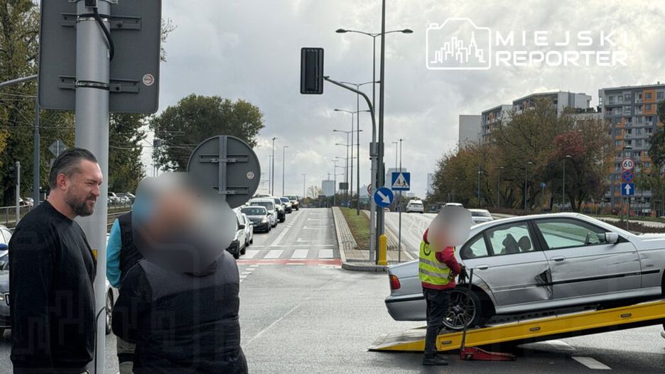 Mężczyzna w czarnym swetrze rozmawia z inną osobą obok drogi, podczas gdy holownik podnosi uszkodzony samochód.