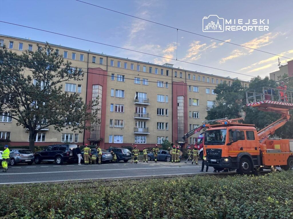 Grupa strażaków pracuje przy usuwaniu skutków zdarzenia drogowego obok wielopiętrowego budynku mieszkalnego.