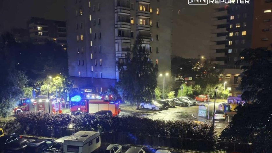 W nocy, w osiedlowym parkingu, straż pożarna z włączonymi syrenami i niebieskimi światłami interweniuje przy budynku mieszkalnym.