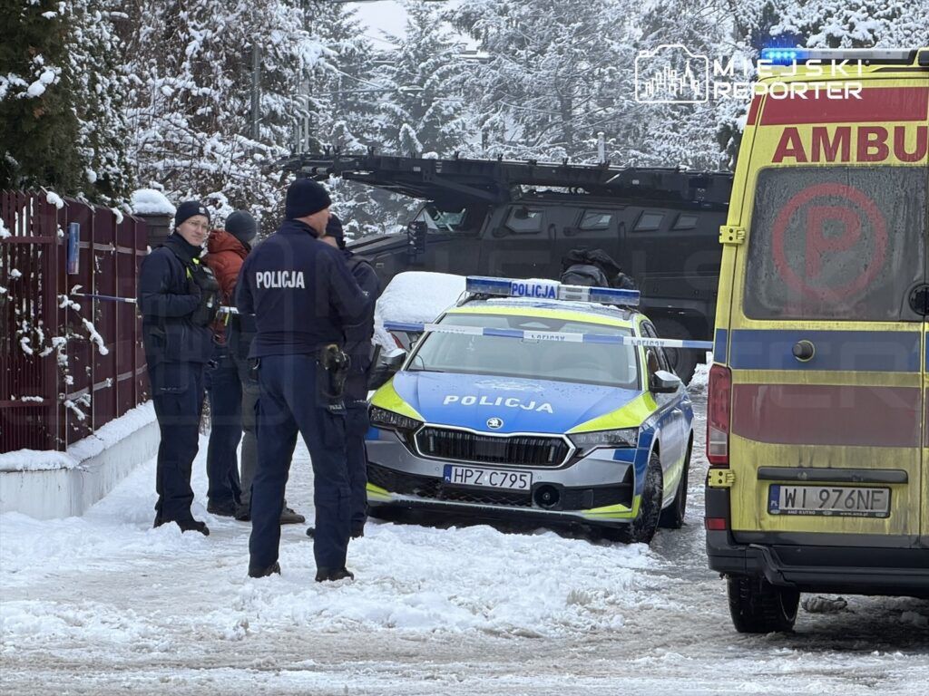 Grupa policjantów w mundurach rozmawia obok radiowozu na zaśnieżonej ulicy, w tle widać ambulans.