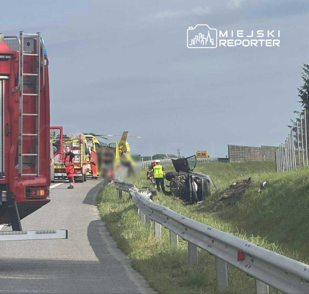 Na drodze leży przewrócone auto, obok pracują ratownicy medyczni i policja, w tle śmigłowiec ratunkowy.