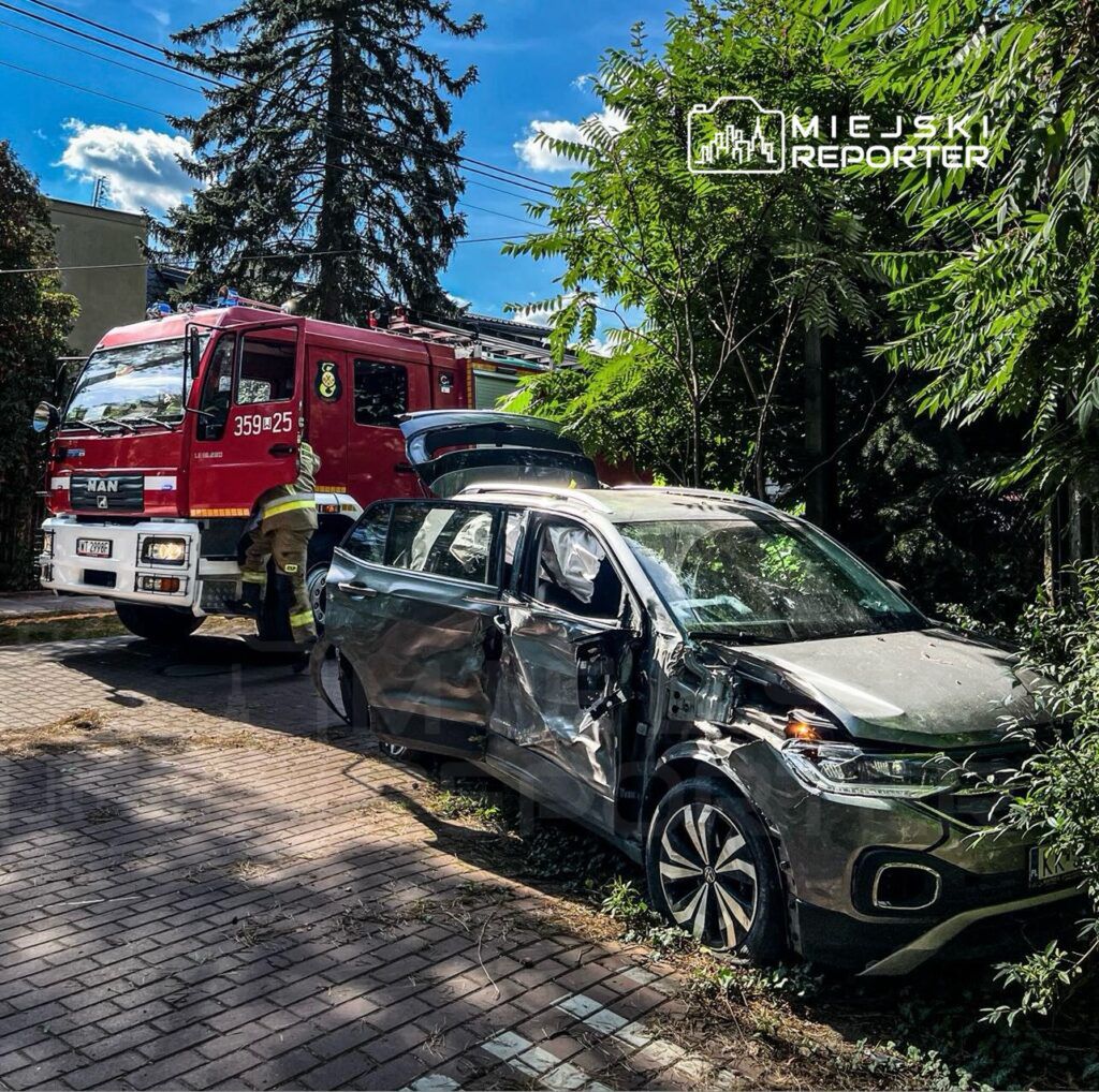 Strażak przy samochodzie osobowym z uszkodzeniami, obok widać wóz strażacki na osiedlowej ulicy otoczonej drzewami.