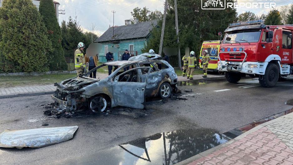 Pojazd pożarowy stoi obok spalonego samochodu, a strażacy prowadzą działania ratunkowe na drodze w terenie zabudowanym.