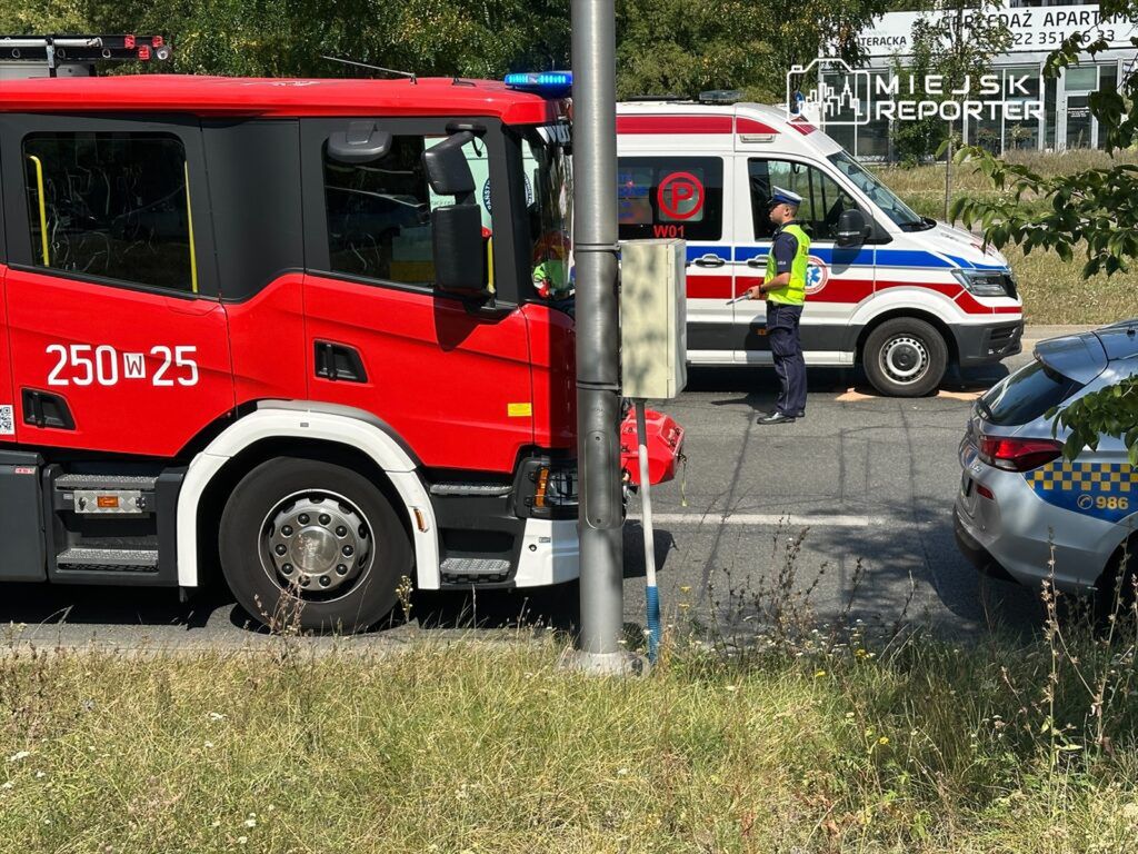 Czerwony wóz strażacki stoi obok karetki pogotowia, a policjant kieruje ruchem na drodze.