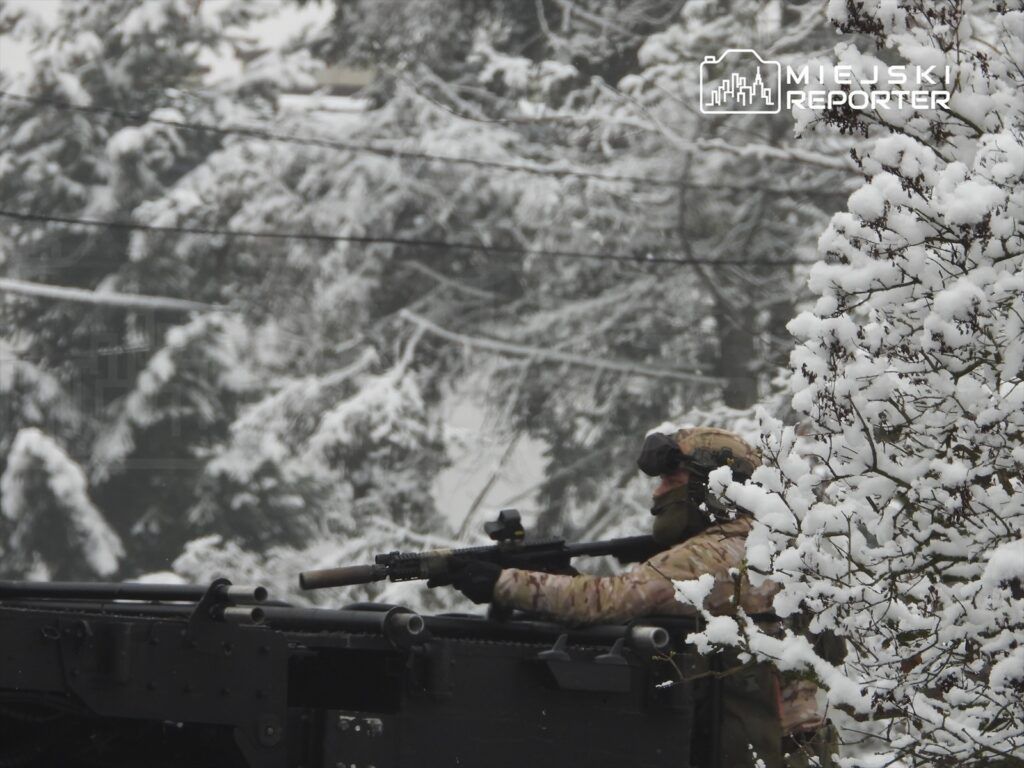 Żołnierz w kamuflażu strzeże terenu z pojazdu opancerzonego, otoczonego zaśnieżonymi drzewami.