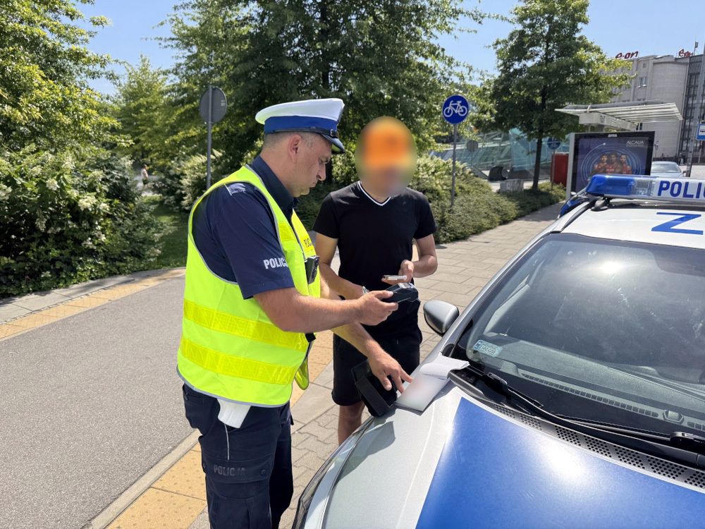 Policjant w kamizelce odblaskowej kontroluje mężczyznę stojącego obok radiowozu na słonecznej ulicy.
