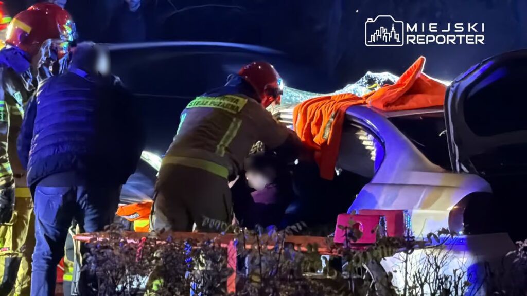 Ratownicy w hełmach pomagają osobie wydobytej z uszkodzonego samochodu, otoczeni przez nocne światła.