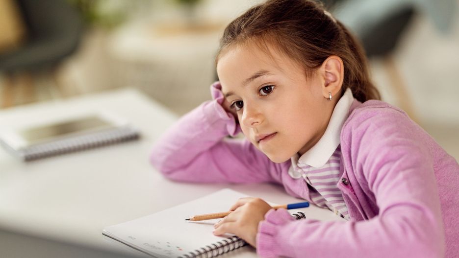 Small girl studying at home and writing in notebook while leaning on a desk.
