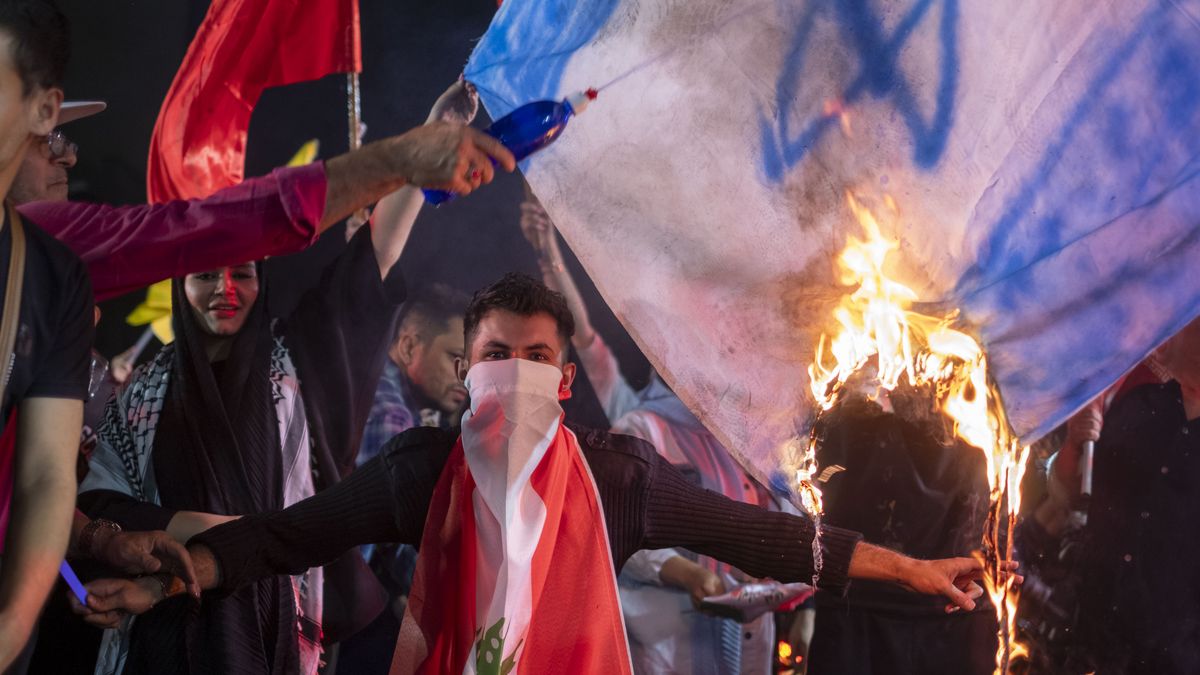 An Iranian man wears a Lebanese flag while protesters burn an Israeli flag during a celebration for Iran's missile attack against Israel in Tehran, Iran, on October 1, 2024. Iran launches hundreds of ballistic missiles at Israel in response to the killings of Hamas Leader Esmail Haniyeh in Tehran and Hassan Nasrallah, the Secretary General of Lebanon's Hezbollah, as well as General Abbas Nilforoushan, a commander in the Islamic Revolutionary Guard Corps' (IRGC) Quds Force, in the suburbs of Beirut, Lebanon. (Photo by Morteza Nikoubazl/NurPhoto via Getty Images)