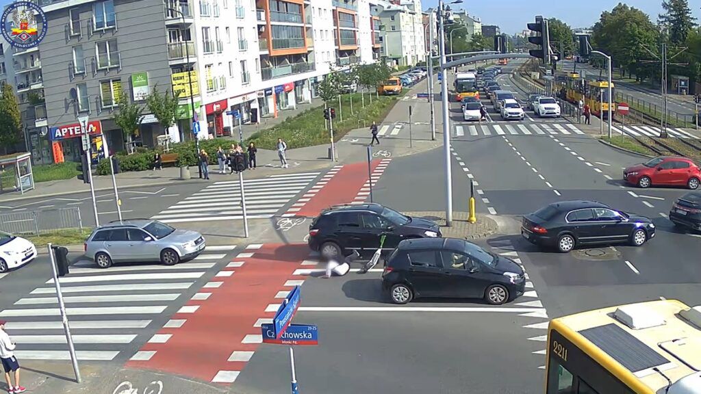 Ruchliwa ulica z przejściem dla pieszych, gdzie samochody zatrzymują się przed sygnalizacją świetlną, a piesi czekają na zielone światło.