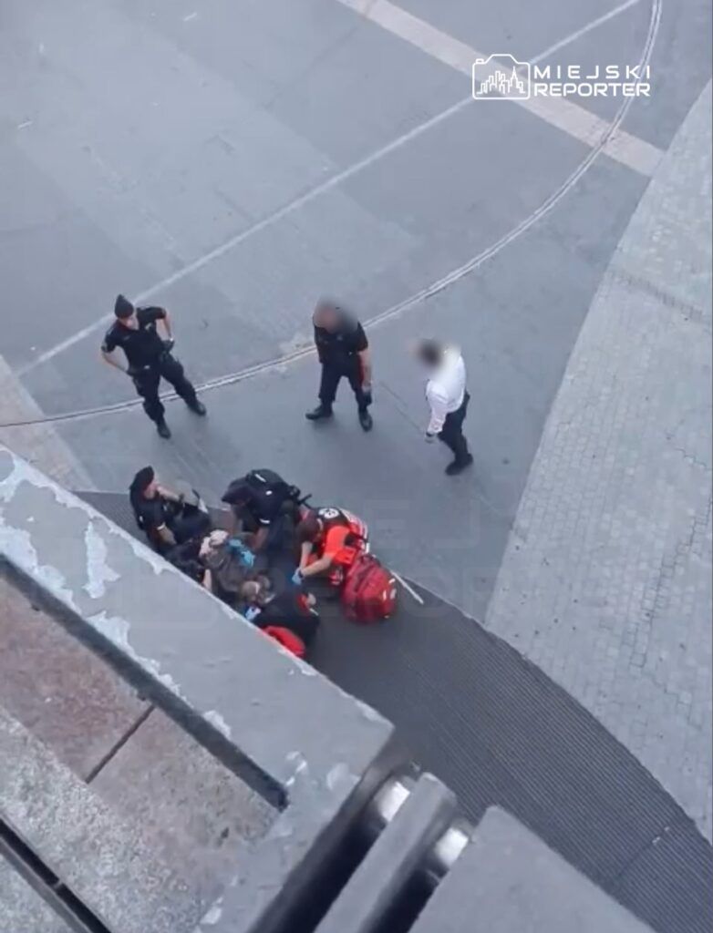 Grupa ratowników i policjantów udziela pomocy osobie leżącej na chodniku w miejskim otoczeniu.