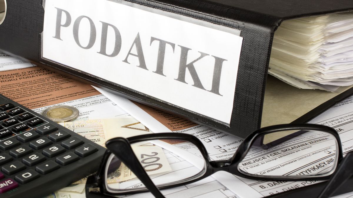 Na zdjęciu formularze podatkowe, na nich leżą okulary, pieniądze i kalkulator. Obok znajduje się segregator z podpisem "podatki"