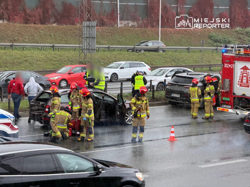 Grupa strażaków udziela pomocy poszkodowanemu na drodze, obok stoi wóz strażacki, a w tle widać ruch uliczny.