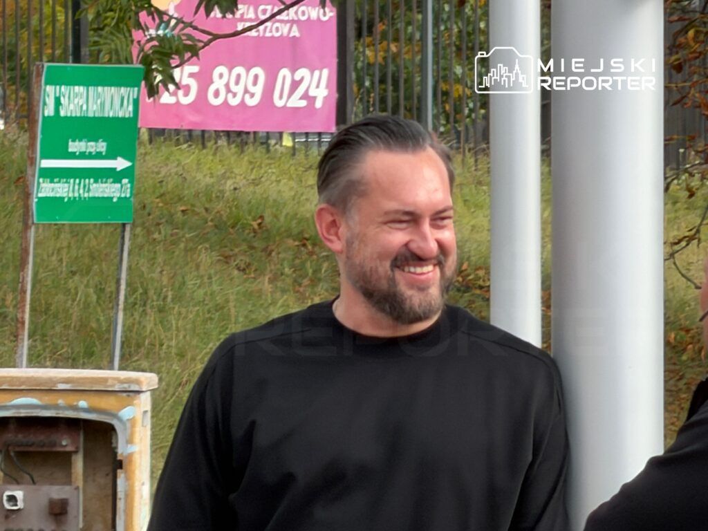 Mężczyzna z brodą uśmiecha się i rozmawia z inną osobą w otoczeniu zieleni i znaków drogowych.
