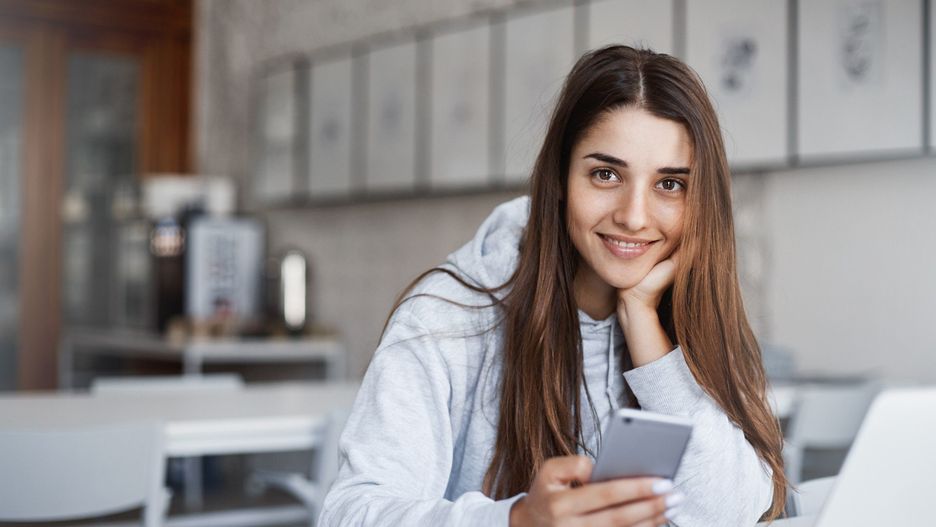 Perfect young woman using smart phone and laptop computer browsing through photos looking at camera smiling rejoicing.