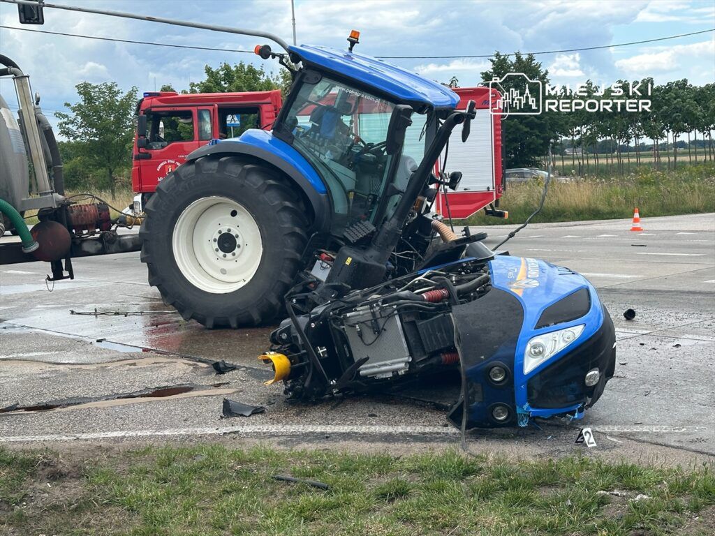Traktor przewrócony na drodze, obok widoczny wóz strażacki, a w tle drzewa i znaki drogowe.