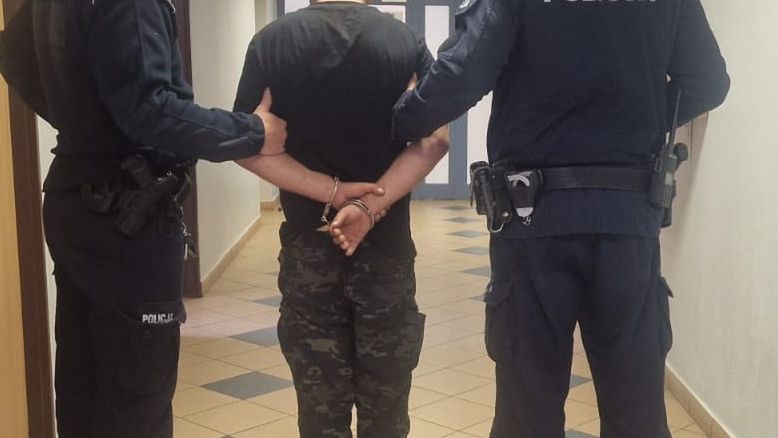 Mężczyzna w kajdankach prowadzony przez dwóch policjantów w korytarzu budynku.