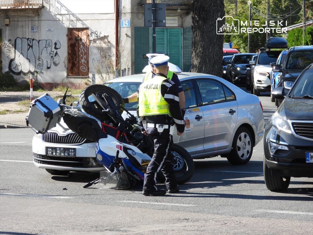 Policjant bada przewrócony motocykl obok zaparkowanego samochodu w miejskim otoczeniu.