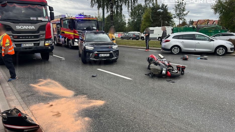 Na jezdni leży przewrócony motocykl, obok widać rozrzucone kaski i ślady piasku, a w tle stoją samochody i straż pożarna.