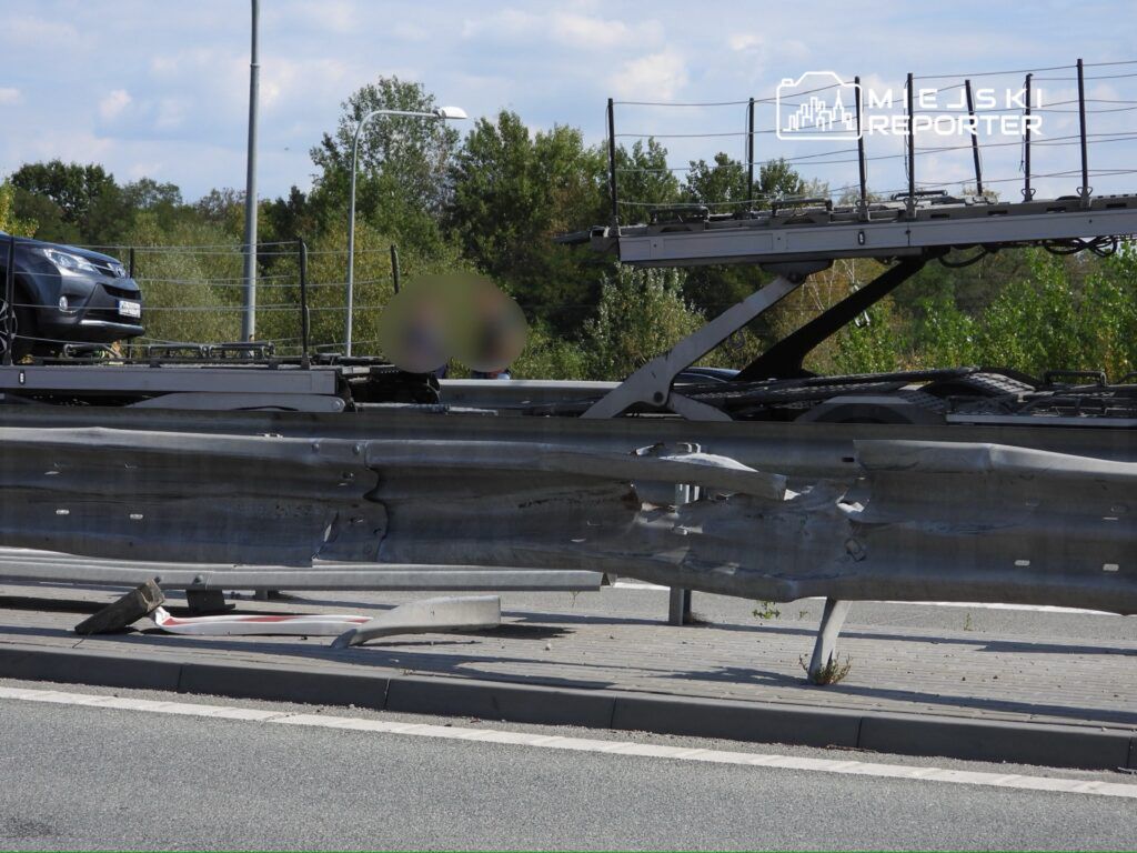 Zniszczona bariera ochronna na drodze, obok stoi uszkodzony pojazd na lawecie. W tle zieleń drzew.