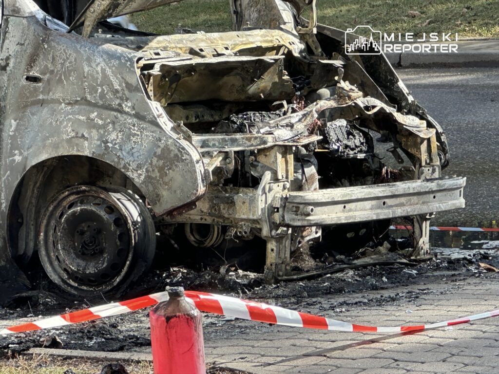 Spalony samochód osobowy z uszkodzoną karoserią i oponami, otoczony taśmą ostrzegawczą, na ulicy.