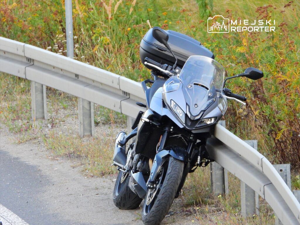 Motocykl w kolorze szarym oparty o barierkę na poboczu drogi, otoczony zielenią.