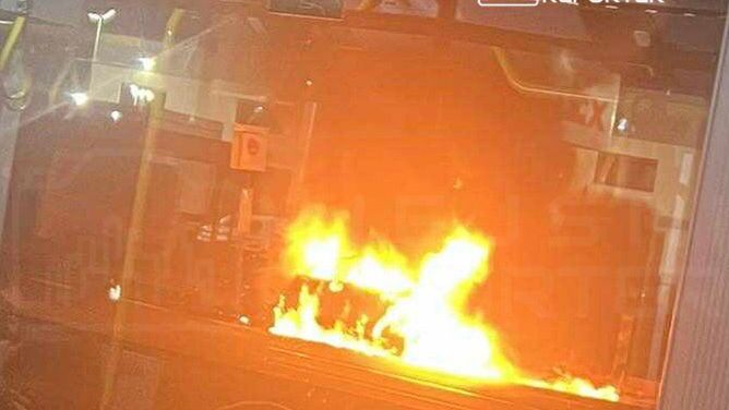 Płonący pojazd komunikacji miejskiej widoczny przez okno, z intensywnym ogniem wydobywającym się z wnętrza.