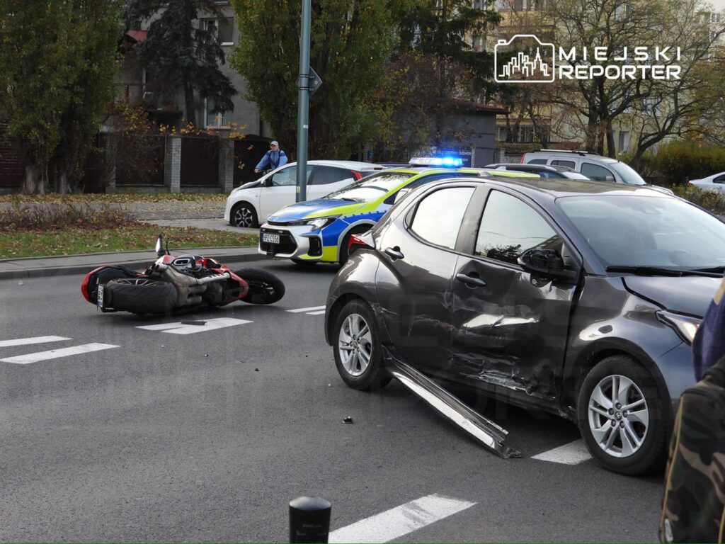 Motocykl leży na boku obok uszkodzonego samochodu osobowego na ulicy, a w tle widać radiowóz policyjny.
