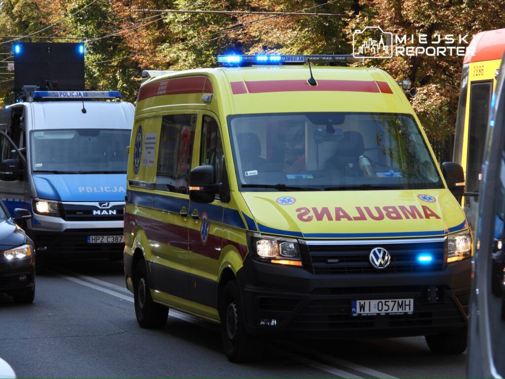 Ambulans z niebieskimi światłami zatrzymany na ulicy, obok policyjny pojazd z logo "POLICJA".