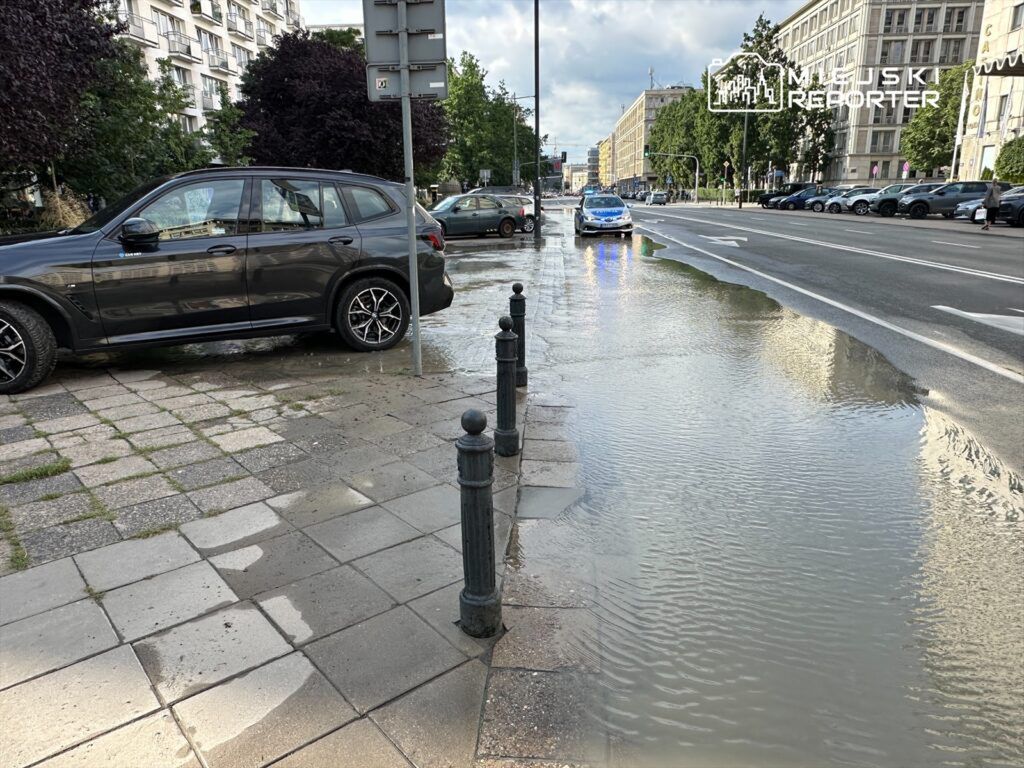 Zalany chodnik obok zaparkowanych samochodów, woda odbija światło wzdłuż ulicy.