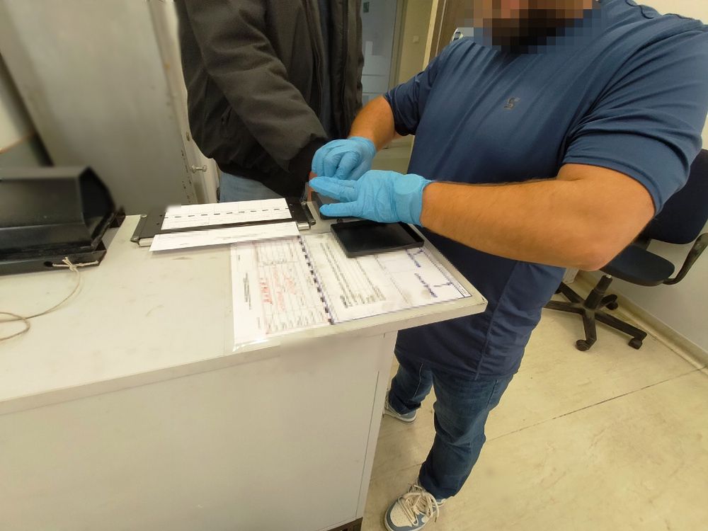 Mężczyzna w niebieskiej koszulce zakłada rękawiczki, przygotowując się do weryfikacji danych przy biurku.