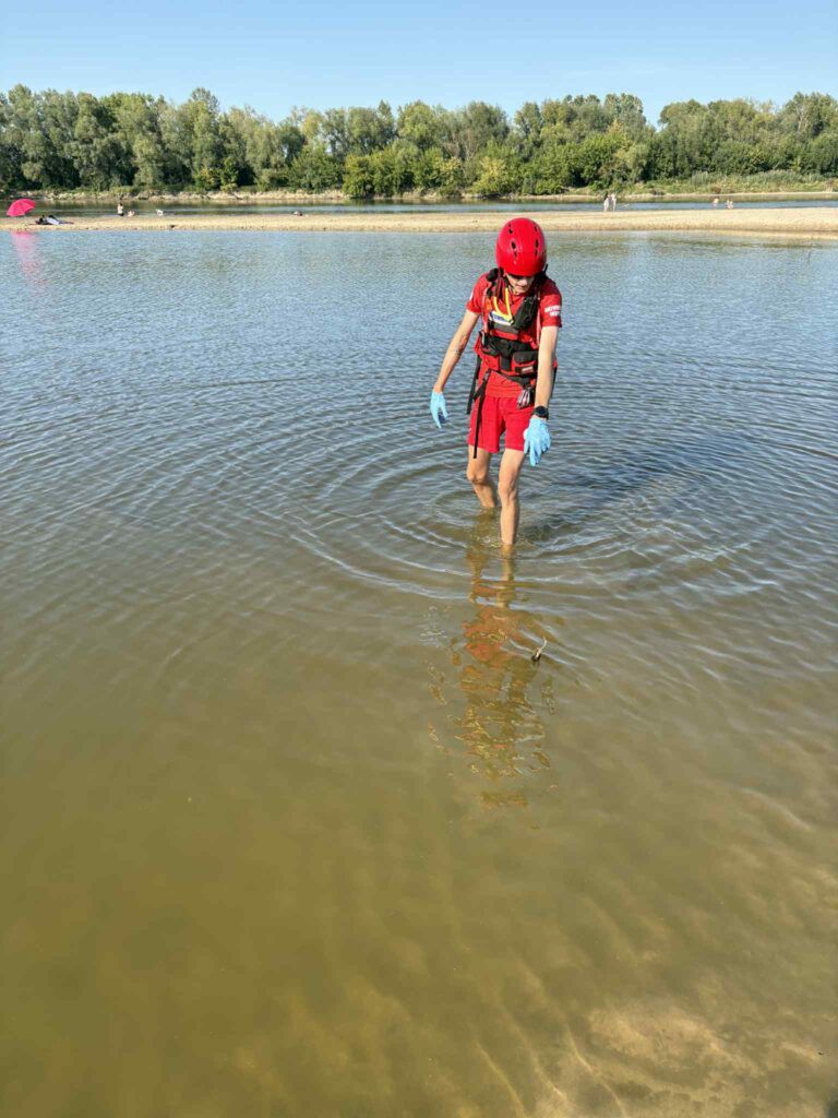 Ratownik wodny w czerwonym stroju i kasku przeszukuje płytką wodę, trzymając w rękach przedmioty. W tle widać plażowiczów.