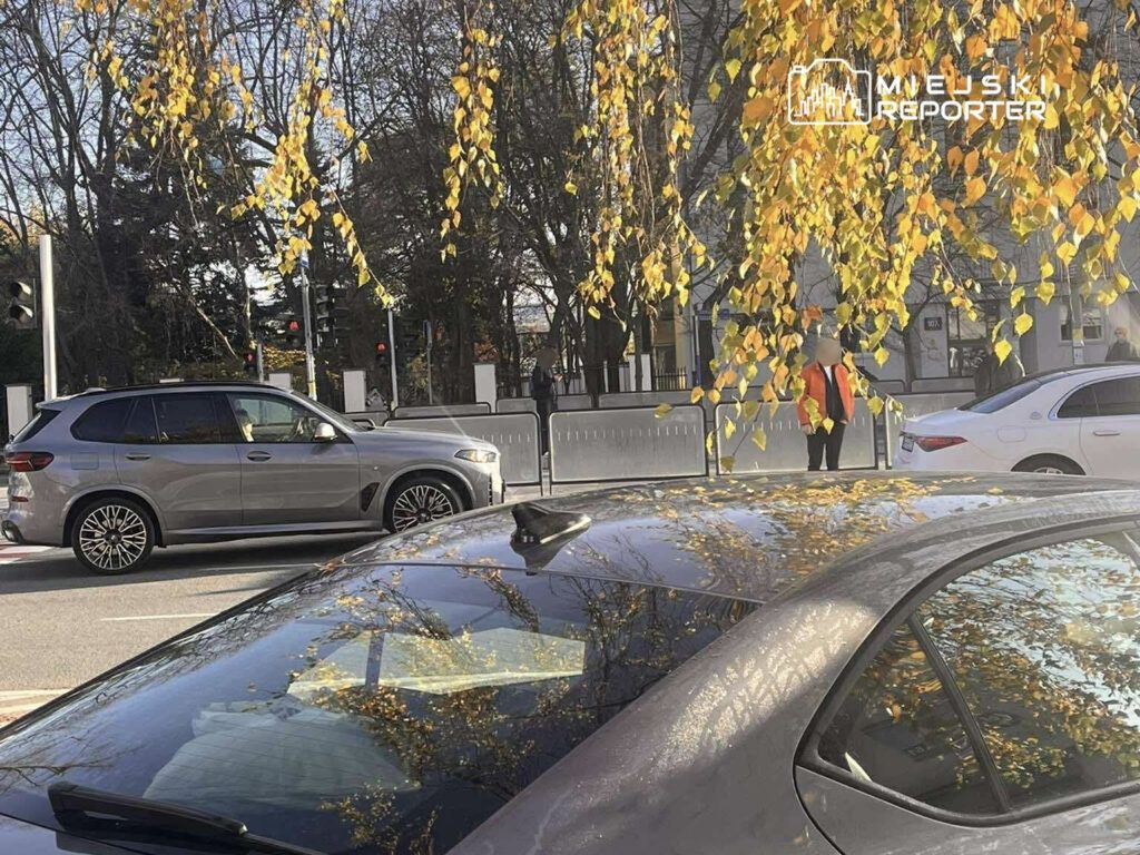Mężczyzna w kamizelce odblaskowej kierujący ruchem na skrzyżowaniu, w tle samochody i drzewa z żółtymi liśćmi.
