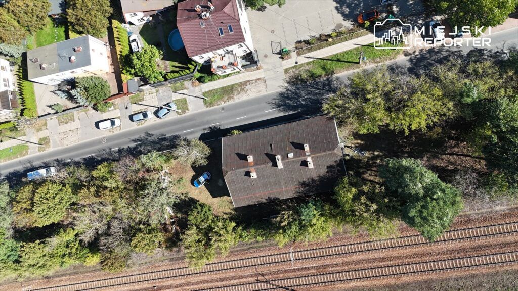 Widok z lotu ptaka na osiedle z domami jednorodzinnymi i torami kolejowymi w tle, otoczone zielenią.