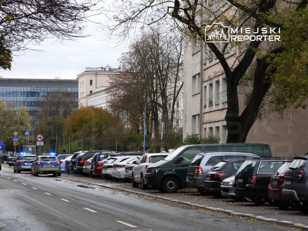 Policyjny radiowóz z włączonymi sygnałami świetlnymi stoi na ulicy obok zaparkowanych samochodów w miejskim otoczeniu.