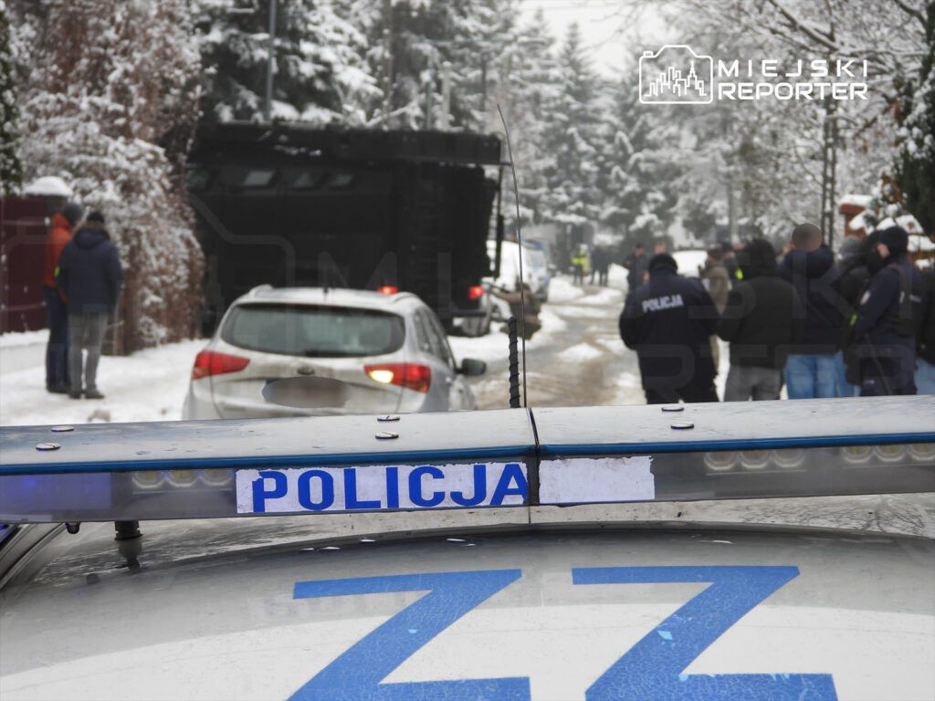 Grupa policjantów w mundurach rozmawia na zaśnieżonej ulicy, obok zaparkowanego samochodu.