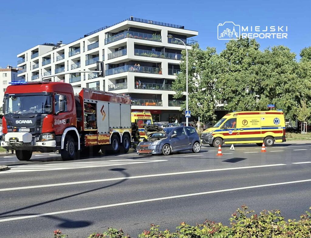Czerwony wóz strażacki i ambulans zatrzymane przy zderzeniu dwóch samochodów na ruchliwej ulicy. W tle nowoczesny budynek mieszkalny.