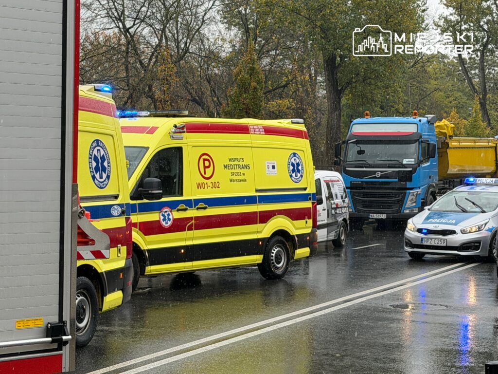 Karetka pogotowia oraz policyjny radiowóz zatrzymane na deszczowej drodze, obok ciężarówki i małego auta.