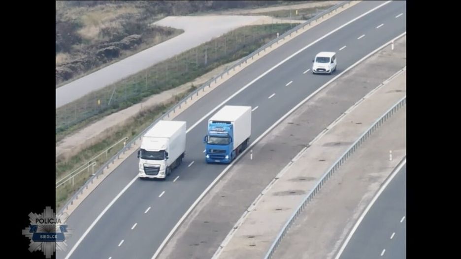 Dwa ciężarówki poruszające się obok siebie na drodze, obok nich mały samochód osobowy. W tle zieleń i pusta przestrzeń.