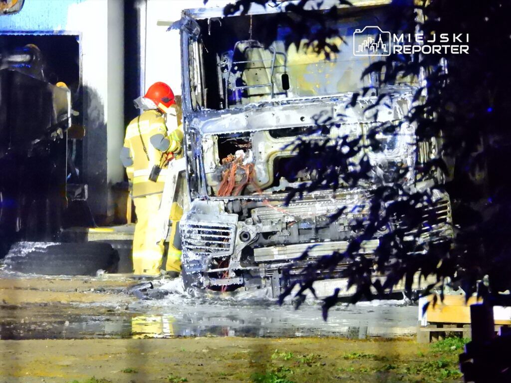 Strażak w hełmie i odzieży ochronnej bada uszkodzony pojazd ciężarowy w nocy, otoczony wodą i sprzętem gaśniczym.