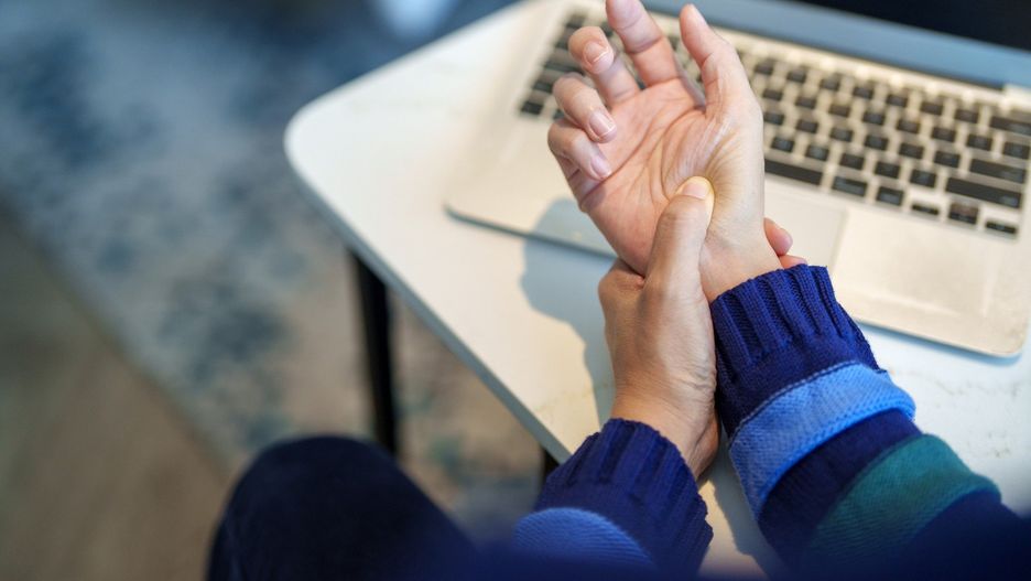 Cropped image of an Asian woman massaging her arthritic hand and wrist after long hours in front of a computer, indicating pain and rheumatism.