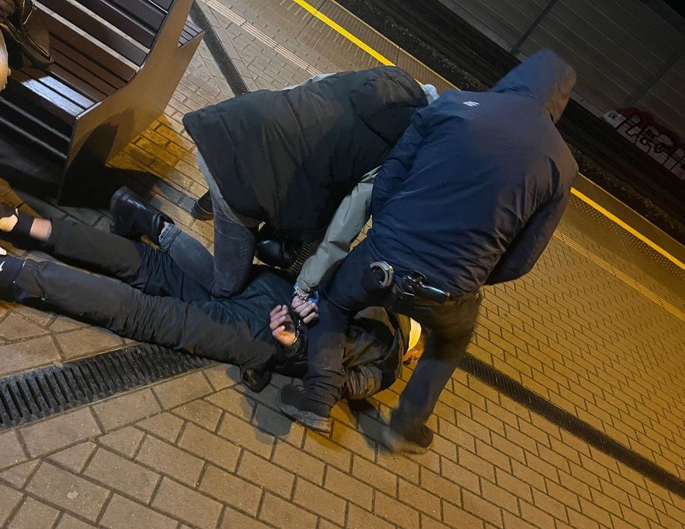 Grupa osób trzyma mężczyznę leżącego na ziemi na peronie, w nocy, w otoczeniu drewnianej ławki i torów kolejowych.