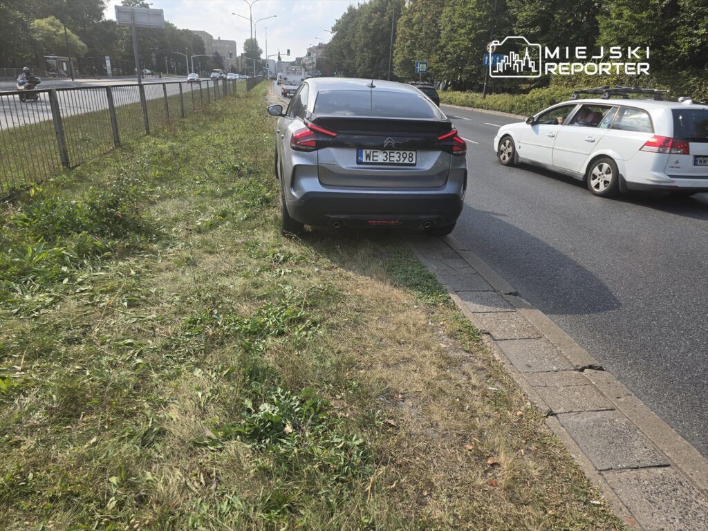 Samochód osobowy zaparkowany na trawie obok ruchliwej ulicy, obok przejścia dla pieszych i metalowego ogrodzenia.
