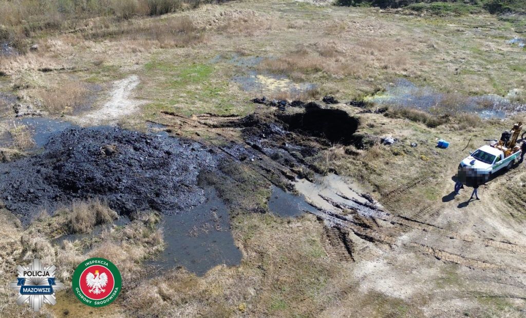 Na zarośniętej łące znajduje się rozległa plama czarnej substancji, otoczona wodą i błotem, obok stoi pojazd służbowy z kilkoma osobami.