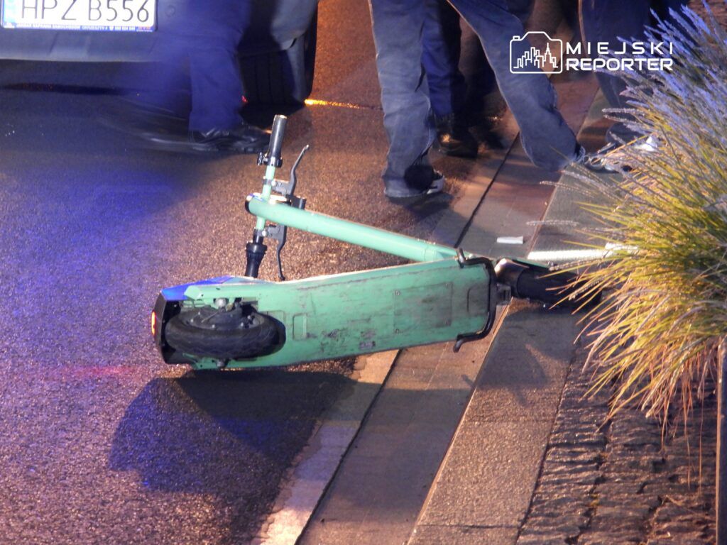 Złamana hulajnoga elektryczna leży na jezdni, obok stojący mężczyzna w ciemnym ubraniu.