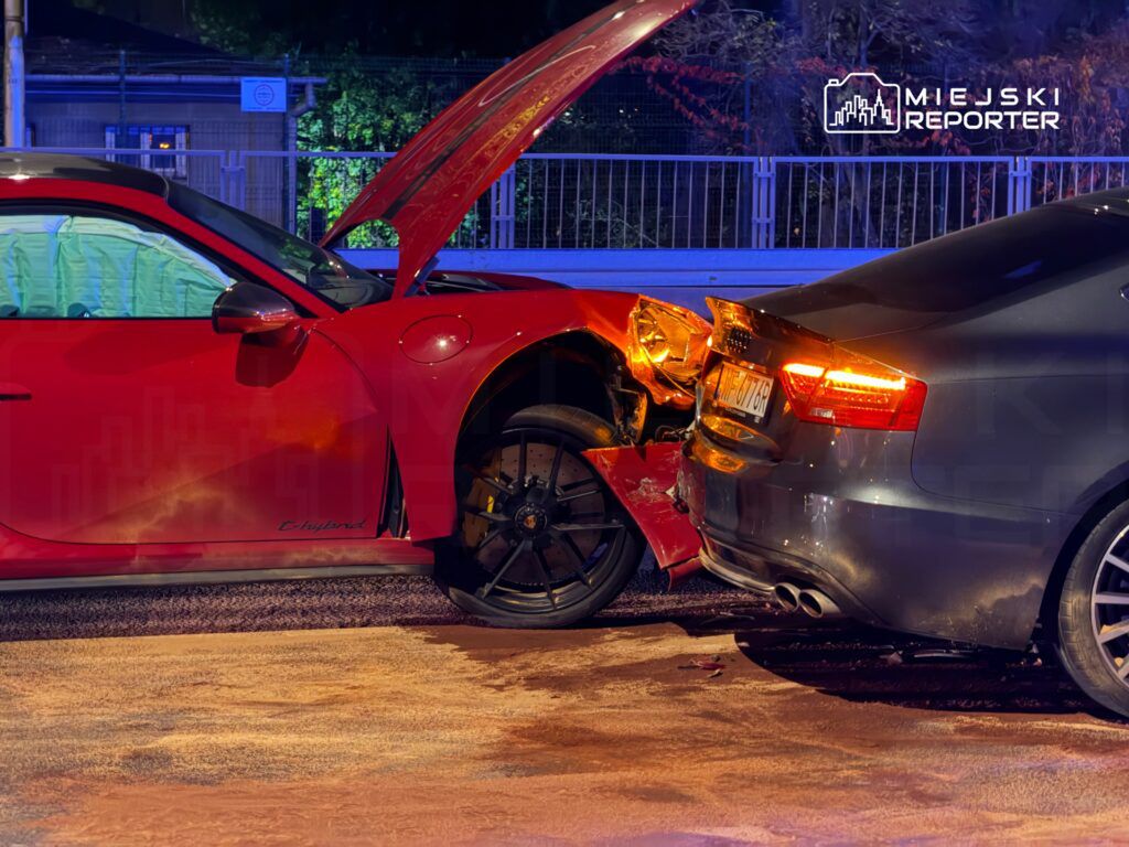 Czerwony samochód sportowy z otwartą maską zderzył się z ciemnoszarym pojazdem na oświetlonej drodze.