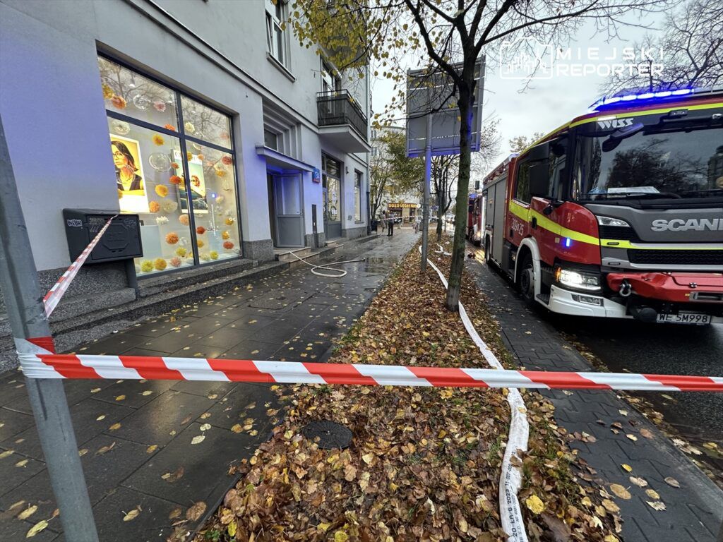 Wzdłuż chodnika znajduje się wąż strażacki, a obok zaparkowany wóz strażacki z włączonymi sygnałami świetlnymi.