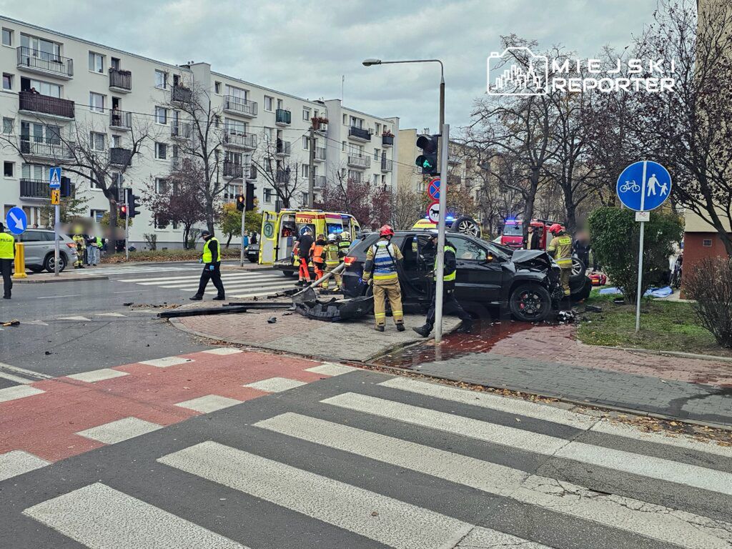 W miejscu wypadku drogowego pracują ratownicy medyczni i strażacy, obok uszkodzonego samochodu na skrzyżowaniu.